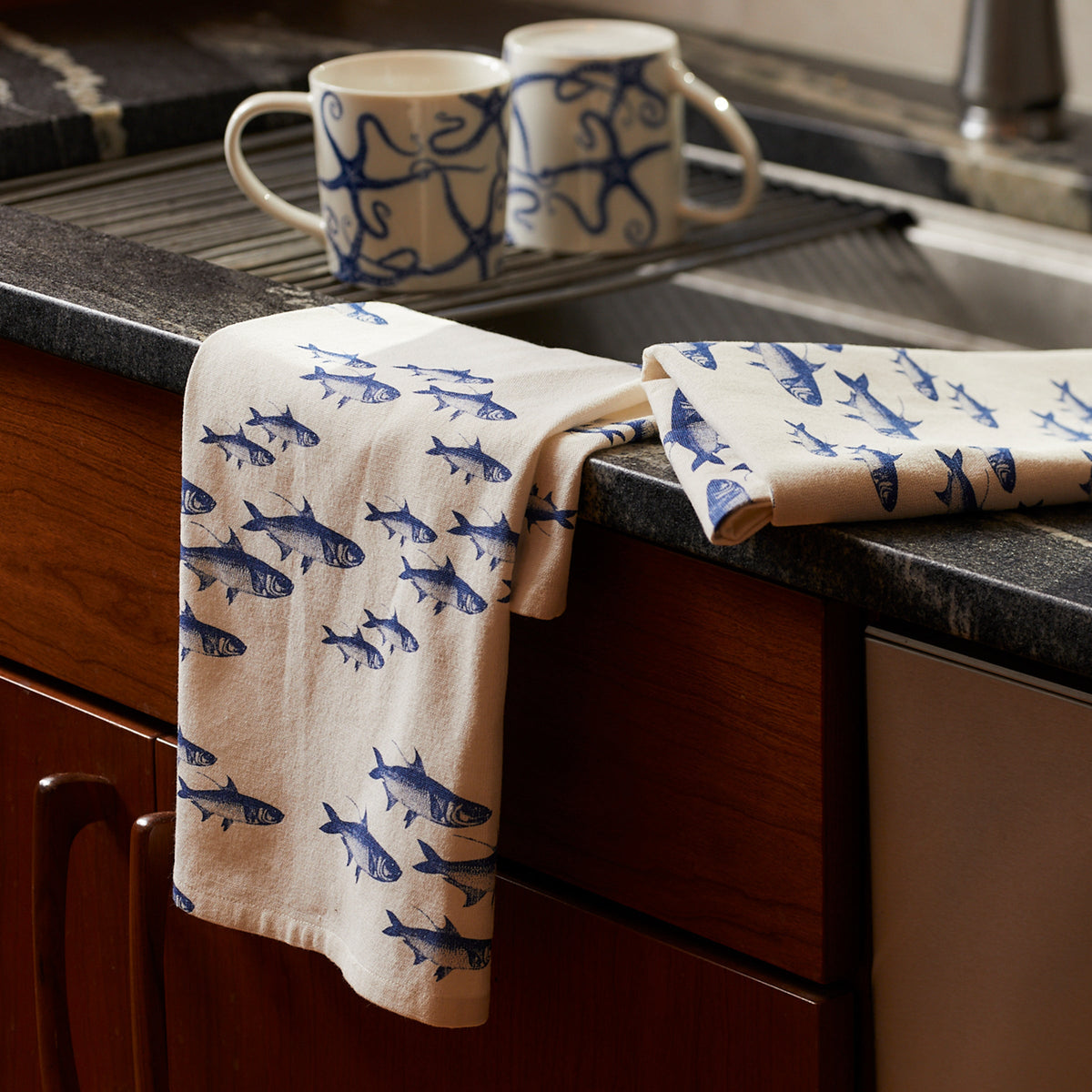 A Caskata School of Fish Kitchen Towel, Set of 2 with blue fish patterns hanging on the edge of a dark countertop, with two matching mugs from a dinnerware collection in the background.