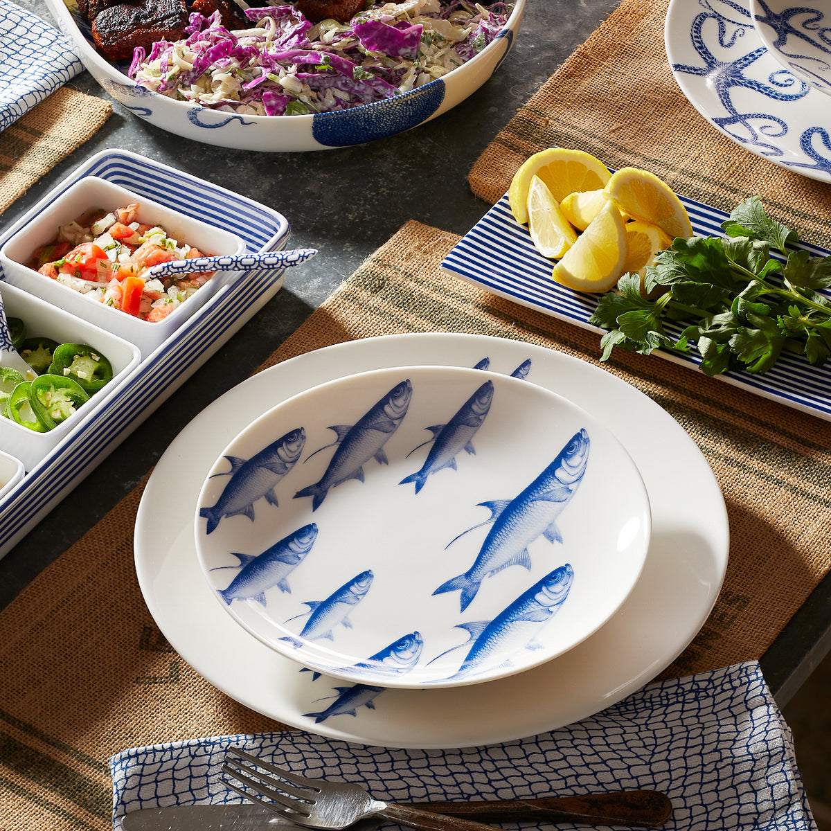 A table setting featuring a School of Fish Coupe Dinner Plate from Caskata Artisanal Home, surrounded by dishes with food, lemons, and herbs.