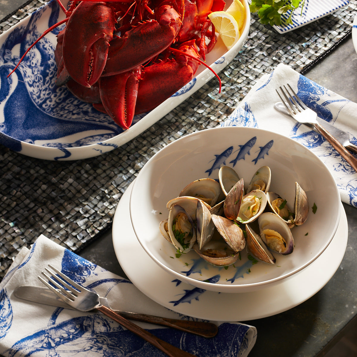 A bowl of clams garnished with herbs sits on a plate next to a dish containing a whole cooked lobster, both pieces from our elegant Caskata Artisanal Home dinnerware collection. Two forks rest on folded napkins beside the plates, ready to make your dining experience delightful.