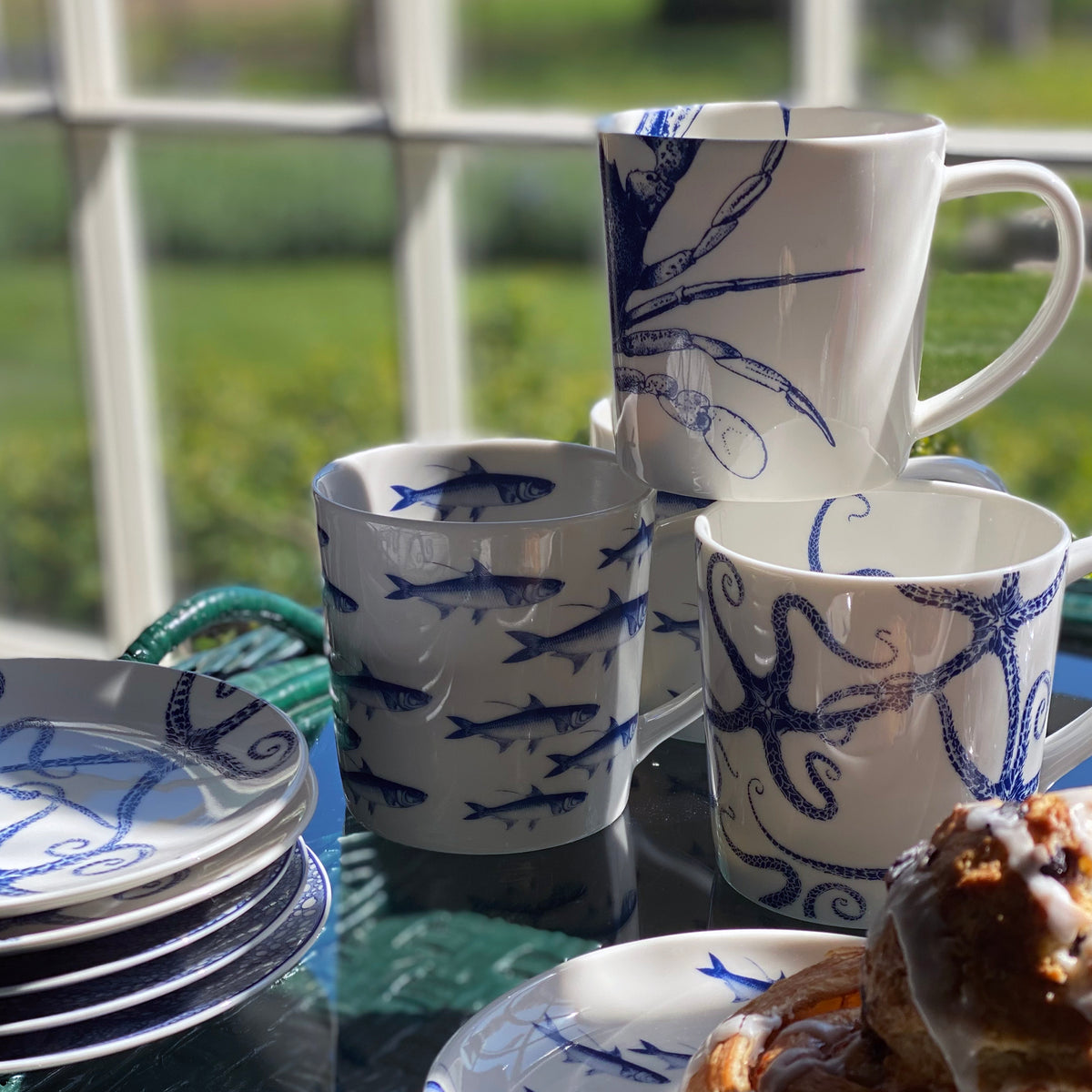 A set of white ceramic plates and Caskata Artisanal Home Starfish Mugs, featuring blue marine life designs, stacked on a glass table by a window. A pastry is partially visible in the foreground. The dinnerware is also dishwasher safe for easy cleaning.