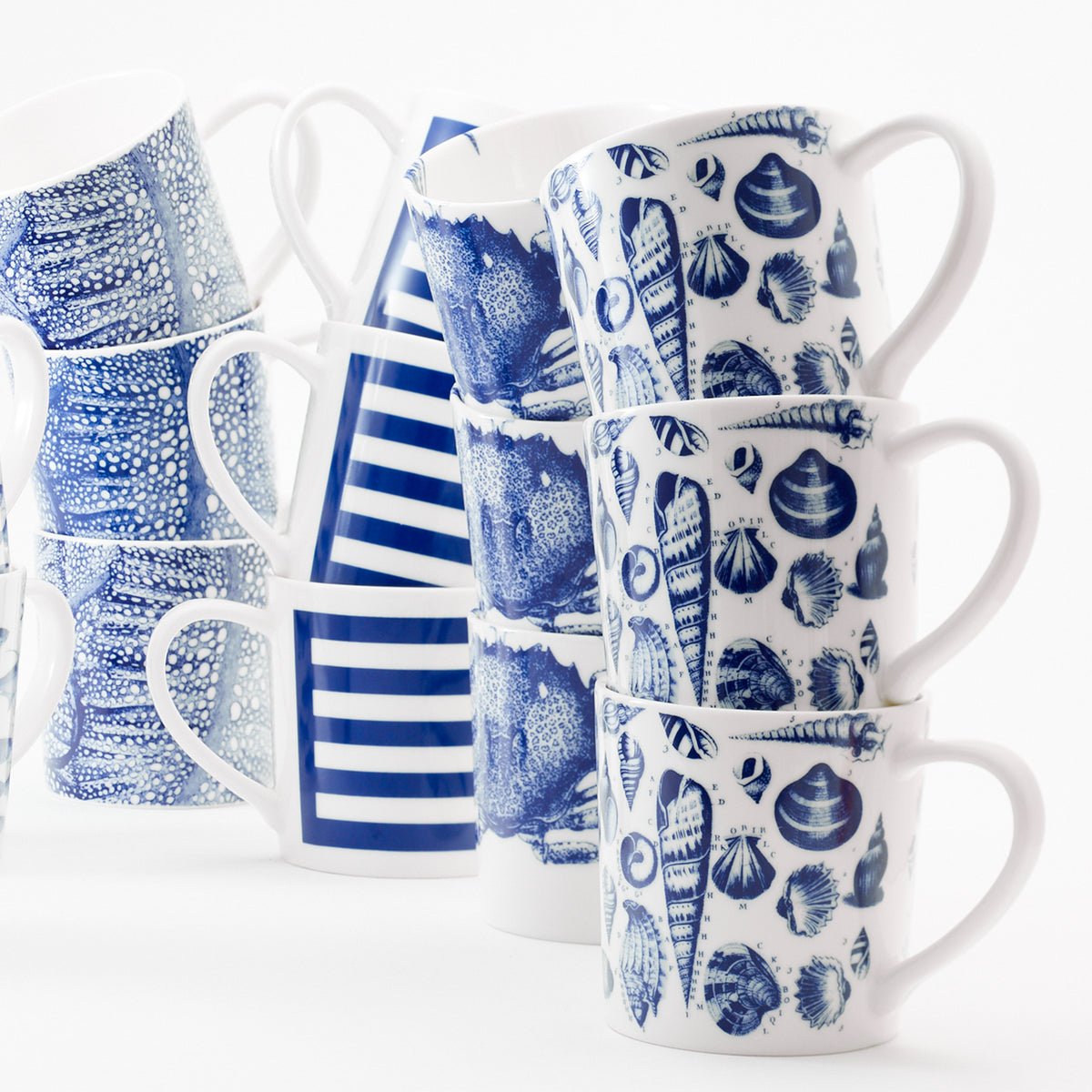A collection of high-fired, beach-inspired Shells Mugs by Caskata Artisanal Home stacked vertically, featuring different patterns such as seashells, stripes, and abstract designs. These blue and white porcelain mugs are also dishwasher safe for easy cleaning.