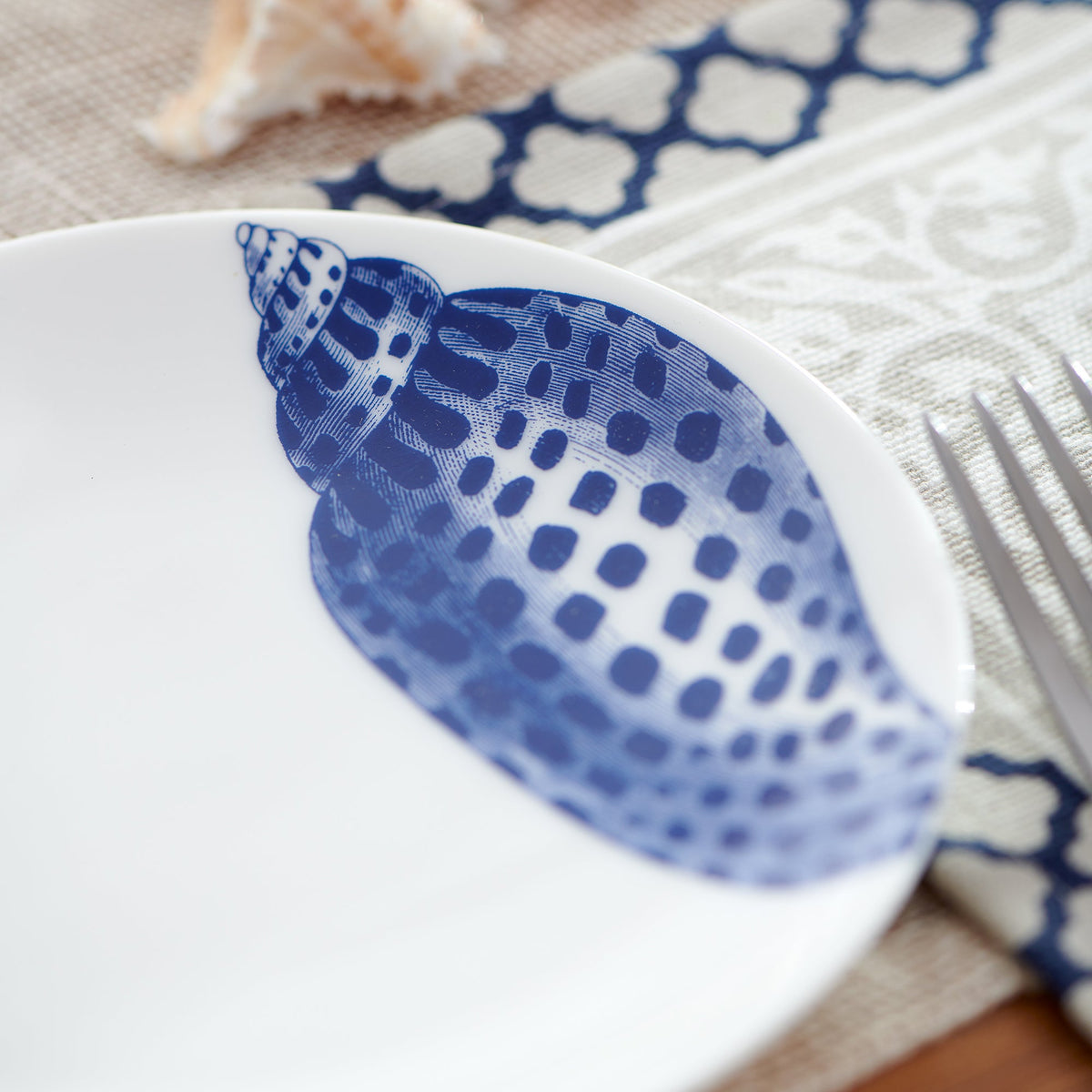 A white plate with a blue seashell design, part of our Caskata Artisanal Home Shells Small Plates, is placed on a patterned tablecloth next to a fork and a small seashell.
