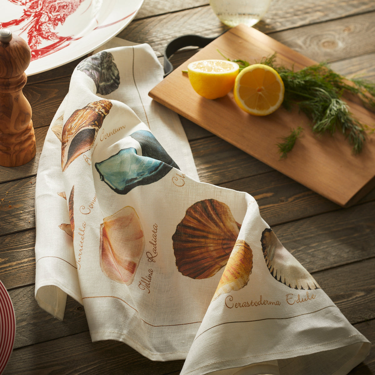 A Shoreline Linen Kitchen Towel adorned with seashells next to a cutting board with lemon and dill from Caskata.
