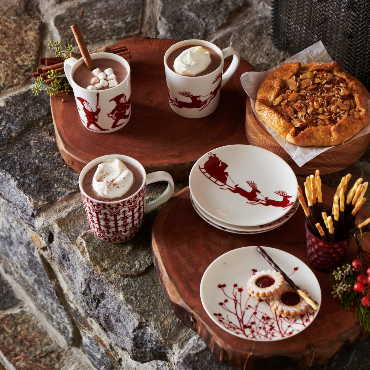 A cozy holiday-themed setup featuring mugs of hot chocolate, Caskata Artisanal Home Sleigh Small Plates with festive designs, a rustic wooden platter with cookies, a pastry, and breadsticks arranged on a stone surface.