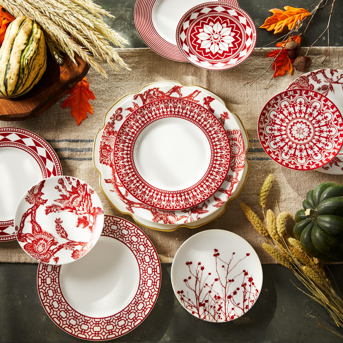 A collection of premium porcelain white plates with red decorative patterns, including Caskata Artisanal Home Newport Garden Gate Crimson Rimmed Salad Plates, arranged on a table with autumn-themed items like dried leaves and gourds.