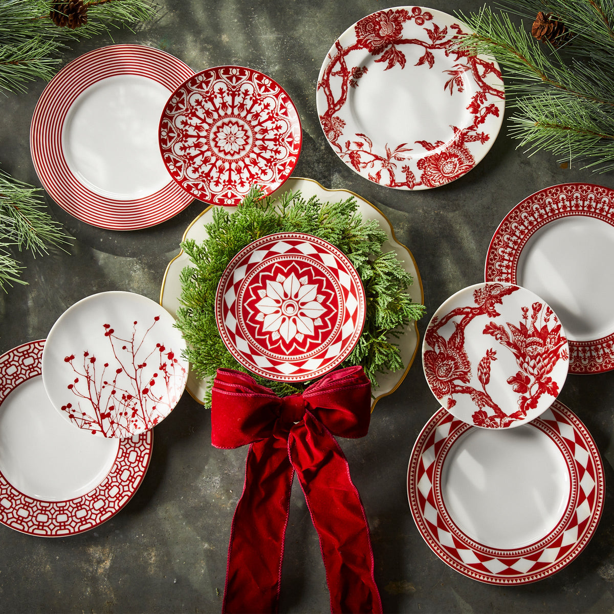 A variety of decorative red and white premium porcelain plates, including elegant **Newport Garden Gate Crimson Rimmed Salad Plates** by **Caskata Artisanal Home**, are arranged in a circular pattern, with a lush wreath featuring a red bow in the center and sprigs of pine framing the display from the corners.