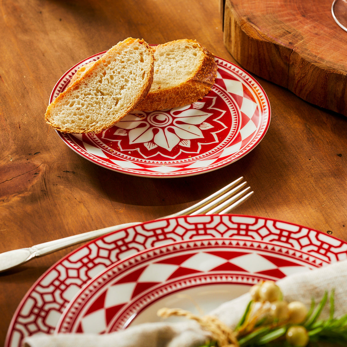 Two slices of bread rest on a Fez Crimson Small Plate from Caskata Artisanal Home, part of an heirloom-quality dinnerware set. A matching larger plate and a fork are placed nearby on the wooden table, creating an entertaining powerhouse for any meal.