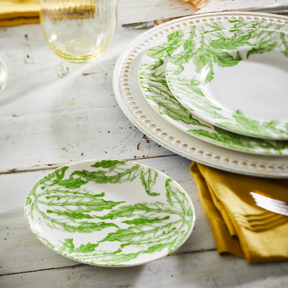 A set of heirloom-quality dinnerware featuring Caskata Artisanal Home&#39;s Freya Small Plates with a green floral pattern arranged on a rustic wooden table, accompanied by a yellow napkin and silver fork.