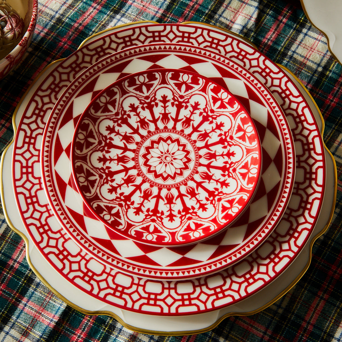 A stack of three high-fired porcelain plates with intricate red and white patterns on a plaid tablecloth. The plates vary in size, with the smallest, resembling a Fez Crimson Rimmed Salad Plate by Caskata Artisanal Home, on top and the largest at the bottom.