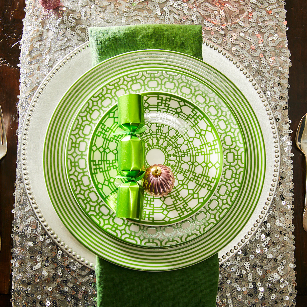 A place setting with a Caskata Artisanal Home Newport Garden Gate Verde Rimmed Salad Plate, green napkin, green cracker, and small pink ornament on a silver sequin tablecloth. This contemporary tableware arrangement evokes a sense of elegance reminiscent of the Newport Garden Gate charm.