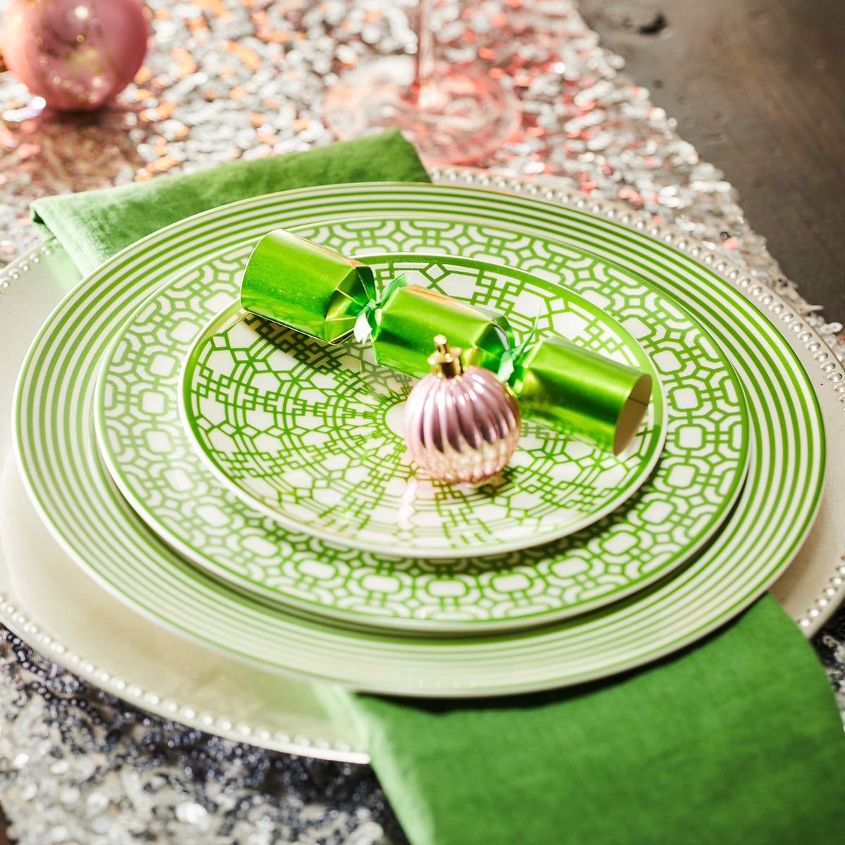 Newport Verde Small Plates by Caskata Artisanal Home, crafted from premium porcelain, with a green napkin, green cracker, and pink ornament set on a textured silver tablecloth.