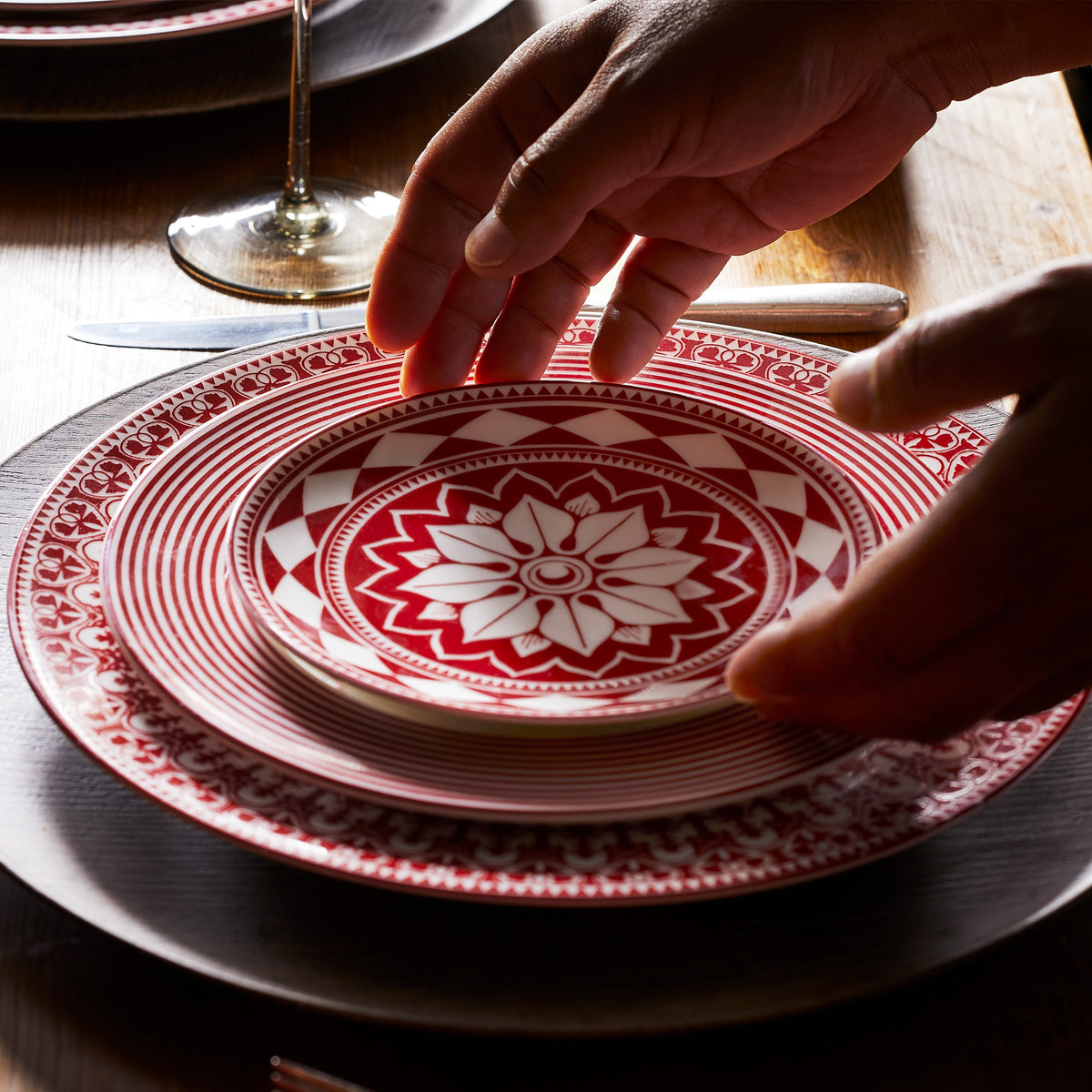 A person places a Fez Crimson Small Plate by Caskata Artisanal Home, showcasing bold geometrics, on top of a larger plate, with a knife and wine glass nearby on a wooden table.