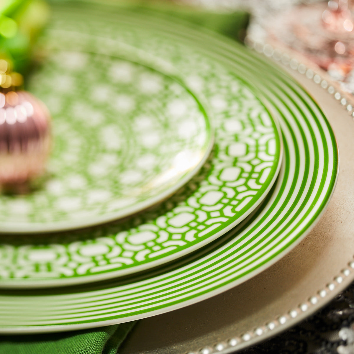 Close-up of patterned green and white Newport Garden Gate Verde Rimmed Salad Plates by Caskata Artisanal Home stacked on a silver charger plate, with a green cloth napkin partially visible underneath.