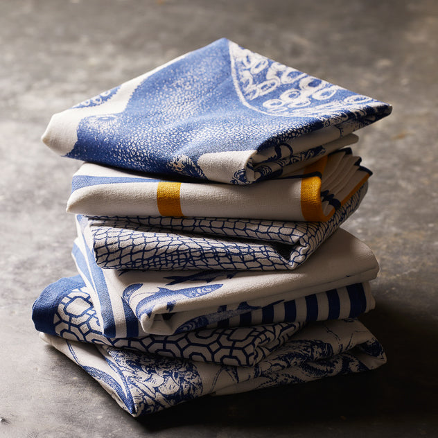 A stack of blue and white table linens, featuring Caskata Lucy Dinner Napkins, Set of 4 with intricate patterns, rests on a gray surface.