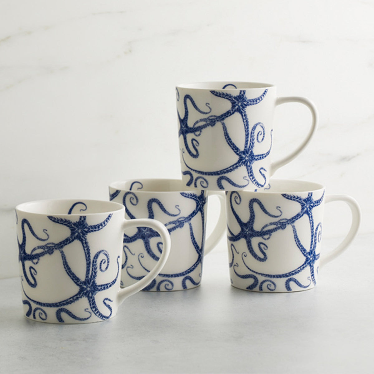 Four high-fired porcelain mugs with blue octopus designs stacked on a gray surface against a white marble background. These dishwasher-safe Starfish Mugs by Caskata Artisanal Home add a touch of marine charm to your kitchen.