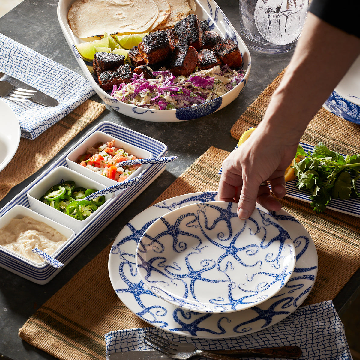 A hand sets a premium porcelain Starfish Coupe Salad Plate by Caskata Artisanal Home on a table with a variety of dishes, including grilled meat, tortillas, sauces, and garnishes. The table is set with brown placemats and silverware.