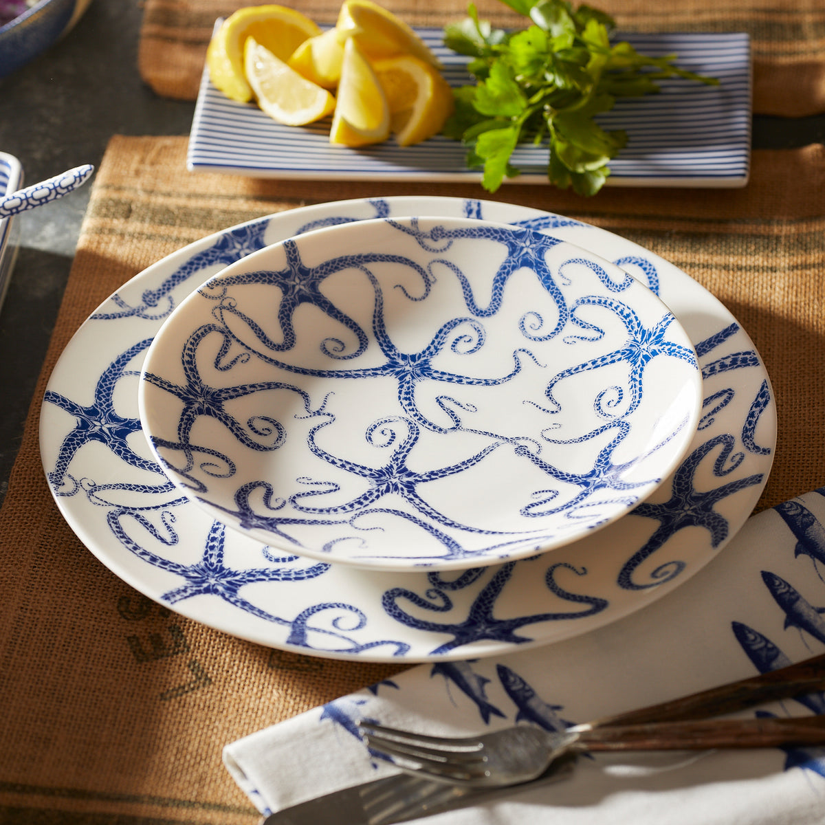 Set of blue and white starfish-patterned dishes on a burlap placemat with a fork and knife on a napkin. A Caskata Artisanal Home Starfish Coupe Salad Plate in the background holds lemon wedges and fresh herbs.