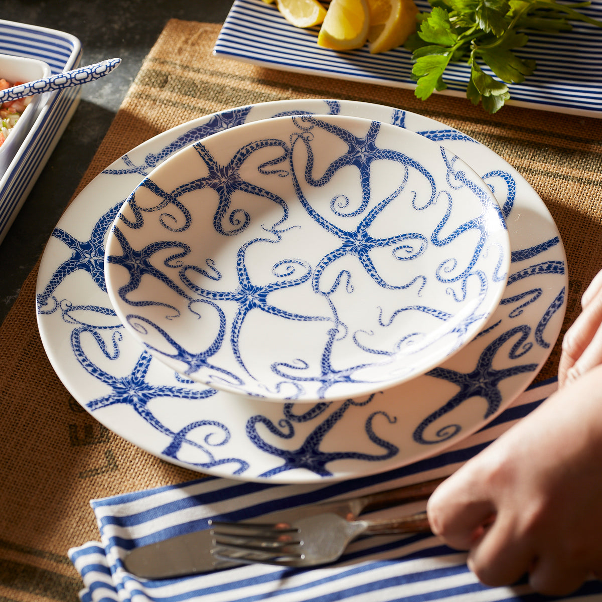 A hand placing a fork and knife onto a striped blue and white napkin next to a Caskata Artisanal Home Starfish Coupe Salad Plate with blue octopus designs on a woven table mat, with herbs and lemon slices in the background.