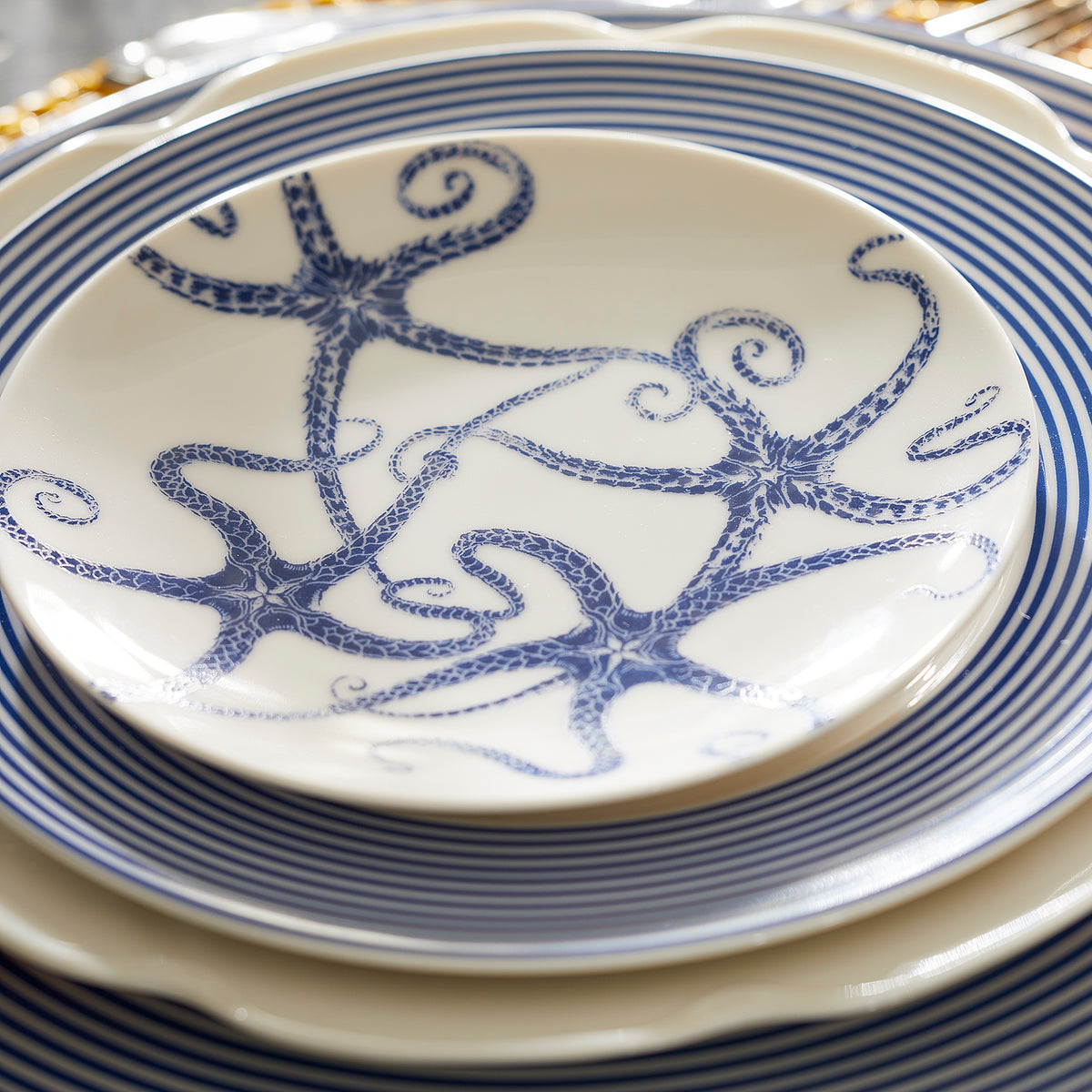 A close-up of stacked heirloom-quality dinnerware in lead-free porcelain with blue nautical designs. The top plate features blue octopus illustrations, while the plates underneath have blue striped patterns. The product shown is Starfish Small Plates by Caskata Artisanal Home.