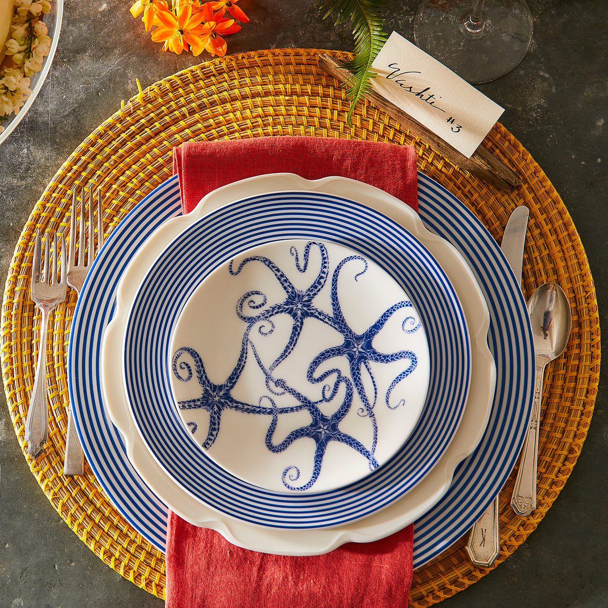 A table setting features a stack of blue and white starfish-themed plates with a red napkin, silver cutlery, and a yellow woven placemat atop a Caskata Artisanal Home Newport Rimmed Charger Plate, accompanied by a name card reading &quot;Yasmin&quot; and an orange flower.
