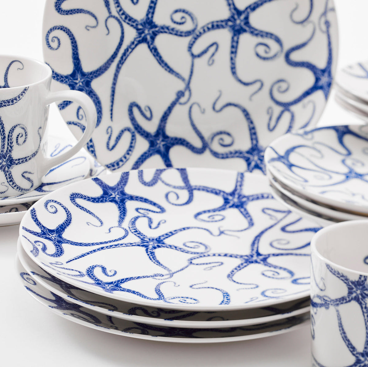 A collection of white premium porcelain dishes, cups, and bowls with blue octopus designs, including a Caskata Artisanal Home Starfish Coupe Salad Plate, arranged on a white surface.