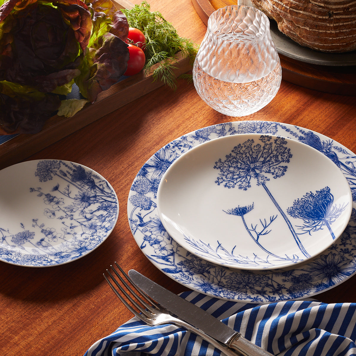 A wooden table set with intricately designed Summer Blues Small Plates by Caskata, a glass of water, a loaf of bread, fresh vegetables, and a striped napkin beside a fork and knife.