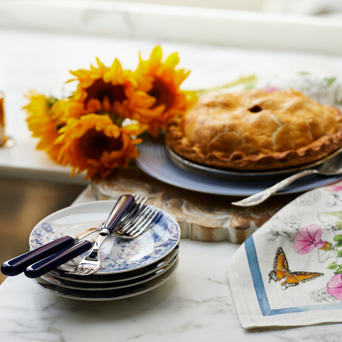 A freshly baked pie on a blue plate, accompanied by sunflowers, a folded napkin with a butterfly design, and a stack of Caskata Summer Blues Small Plates with utensils on a marble counter.