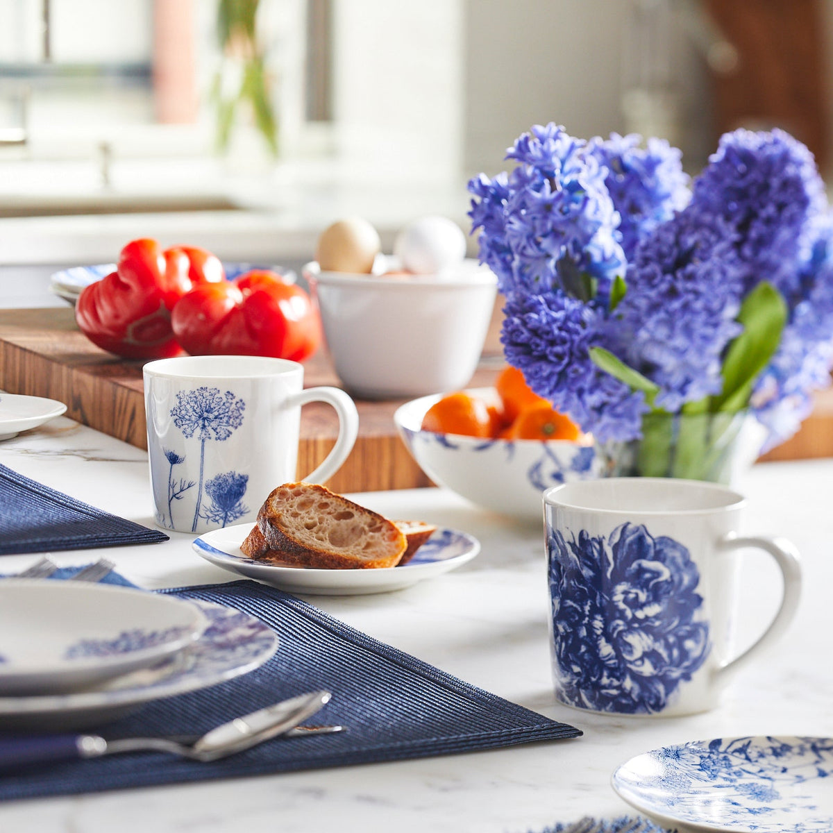 Bright kitchen table setting featuring Caskata Artisanal Home premium porcelain blue and white dinnerware, Peony mugs, fresh fruit, bread, and a vibrant bouquet of blue flowers.