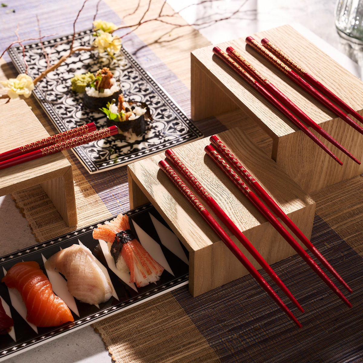 A set of red chopsticks arranged on wooden stands next to plates of assorted sushi and rolls, placed on a Caskata Casablanca Large Sushi Tray atop a bamboo mat.