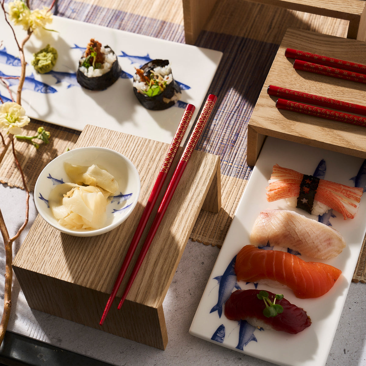 A display of assorted sushi including nigiri and rolls on a **Caskata School of Fish Large Sushi Tray**, ginger slices in a dish, two sets of red chopsticks, and a branch with flowers on a wooden and bamboo mat background.