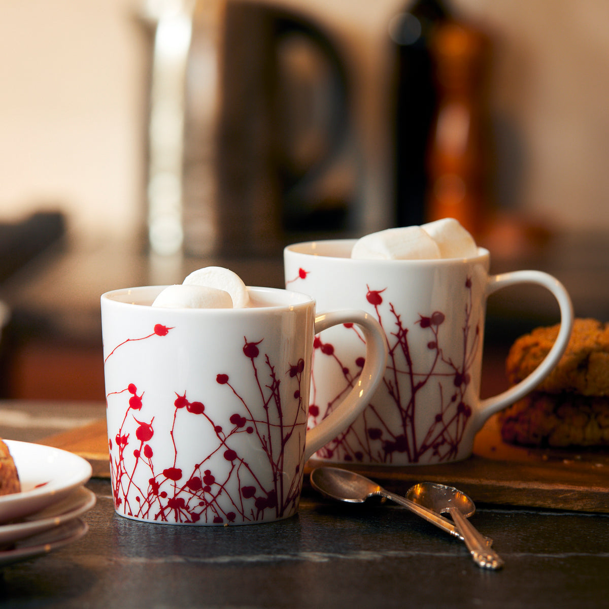 Two generously sized Winterberries Mugs by Caskata Artisanal Home, filled with hot drinks topped with marshmallows, are on a creamy white porcelain kitchen counter. Spoons lie in front, and cookies rest nearby. Blurred background shows kitchen items.