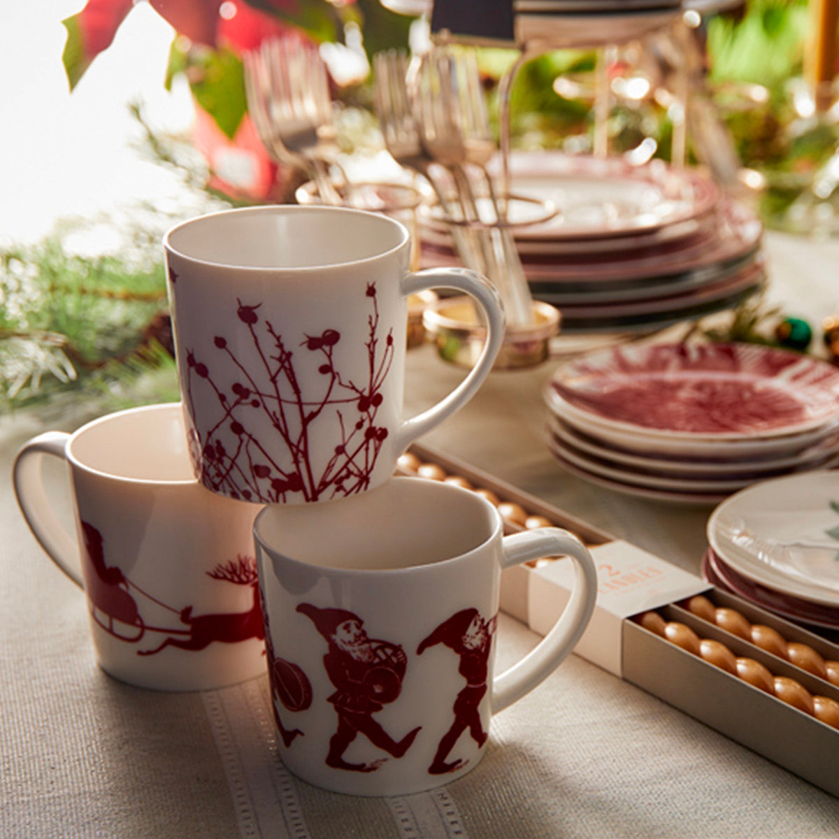 Three Elves Mugs from Caskata Artisanal Home with red holiday designs are stacked on a decorated table, surrounded by an assortment of plates, flatware, and festive accents. This holiday collection exudes charm with hints of playful elves adding a touch of whimsy to the scene.