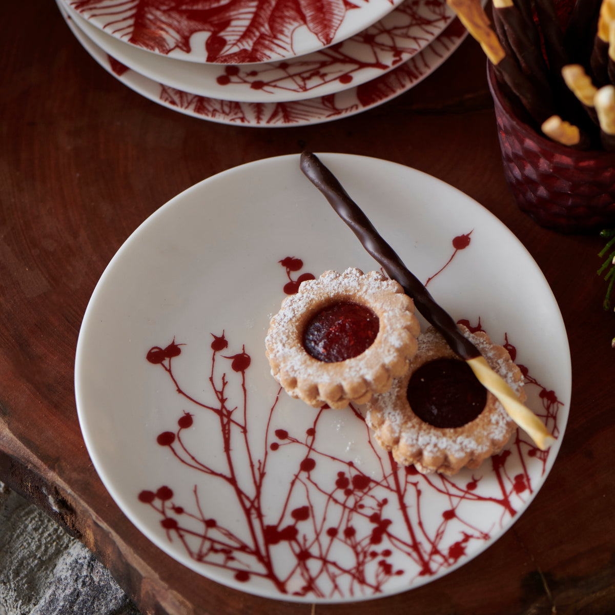 A white plate adorned with Winter berries and a red floral design holds two round cookies with jam centers and a single chocolate-dipped stick cookie rests on a **Caskata Artisanal Home Winterberries Small Plate**.