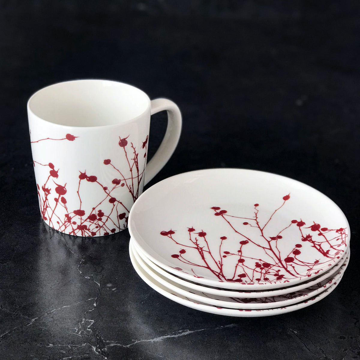A white mug and a stack of three Winterberries Small Plates from Caskata Artisanal Home, crafted from heirloom-quality porcelain, are decorated with a red floral design resembling winter berries on a dark surface.