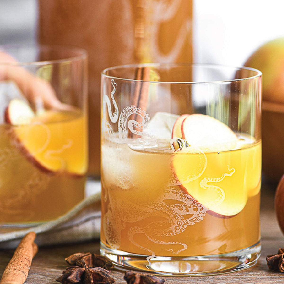 Two glasses of a light brown drink with ice, garnished with apple slices and cinnamon sticks. The Lucy Rocks Glasses by Caskata Artisanal Home boast an intricate design. Star anise and a whole apple are in the background, adding to the autumnal charm.