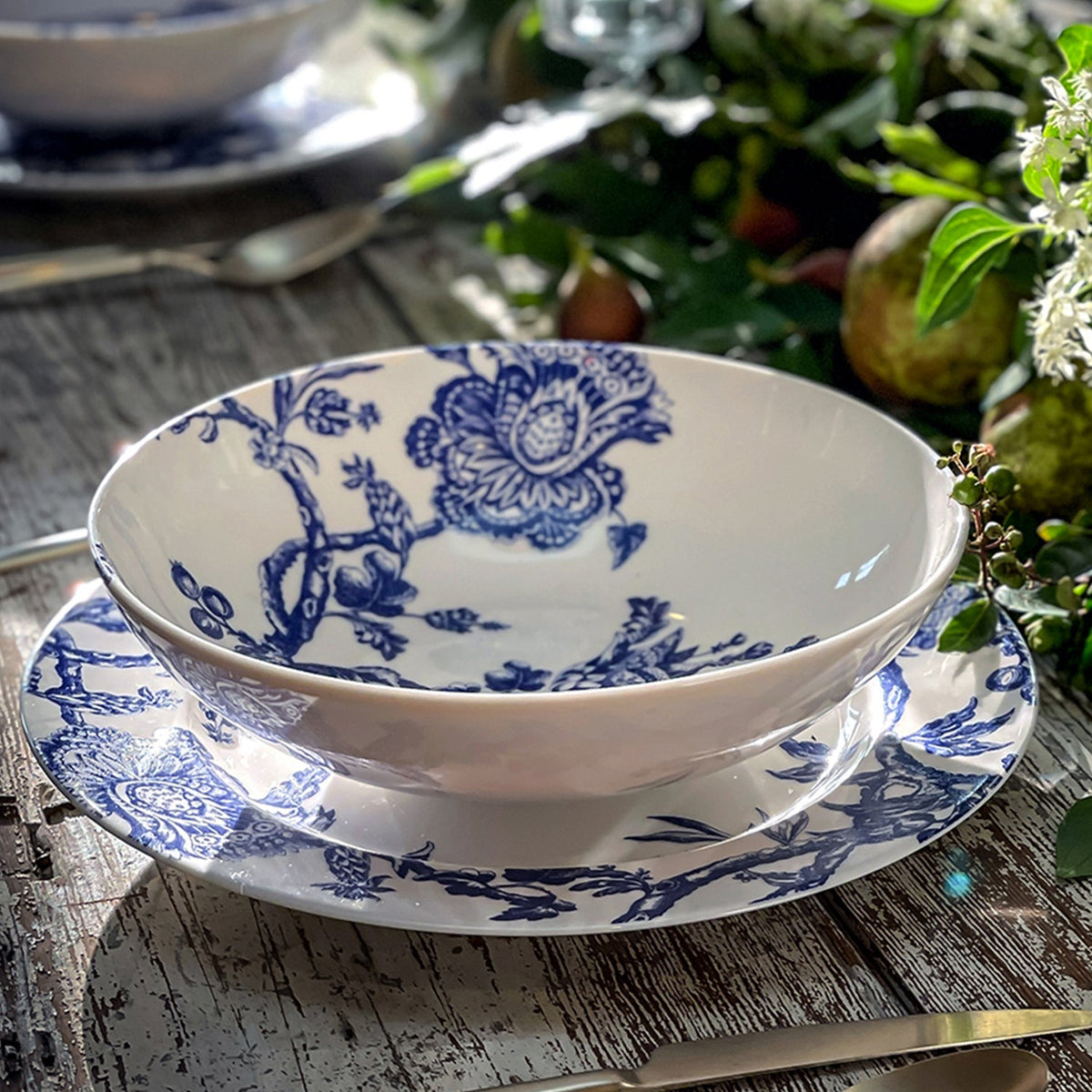 A generous Arcadia Entrée Bowl by Caskata Artisanal Home with blue floral patterns, part of the premium porcelain dinnerware collection inspired by the Williamsburg Foundation, rests on a matching plate on a rustic wooden table with greenery and fruit decorations in the background.