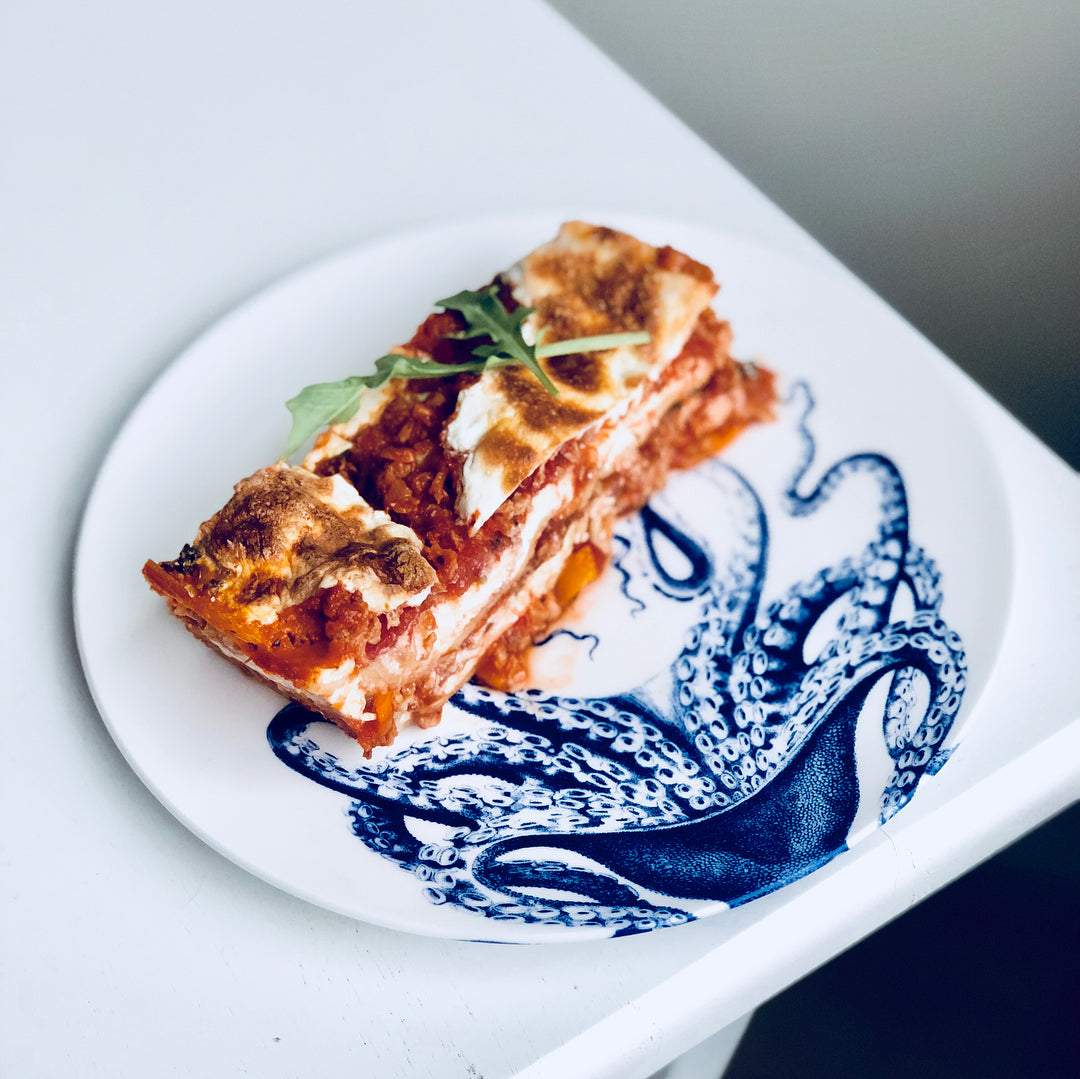 A piece of lasagna topped with arugula is served on a white plate with a blue octopus design, crafted from creamy white premium porcelain. Its contemporary shape adds elegance to your meal, and the Lucy Coupe Salad Plate by Caskata Artisanal Home is both dishwasher and microwave safe for convenience.