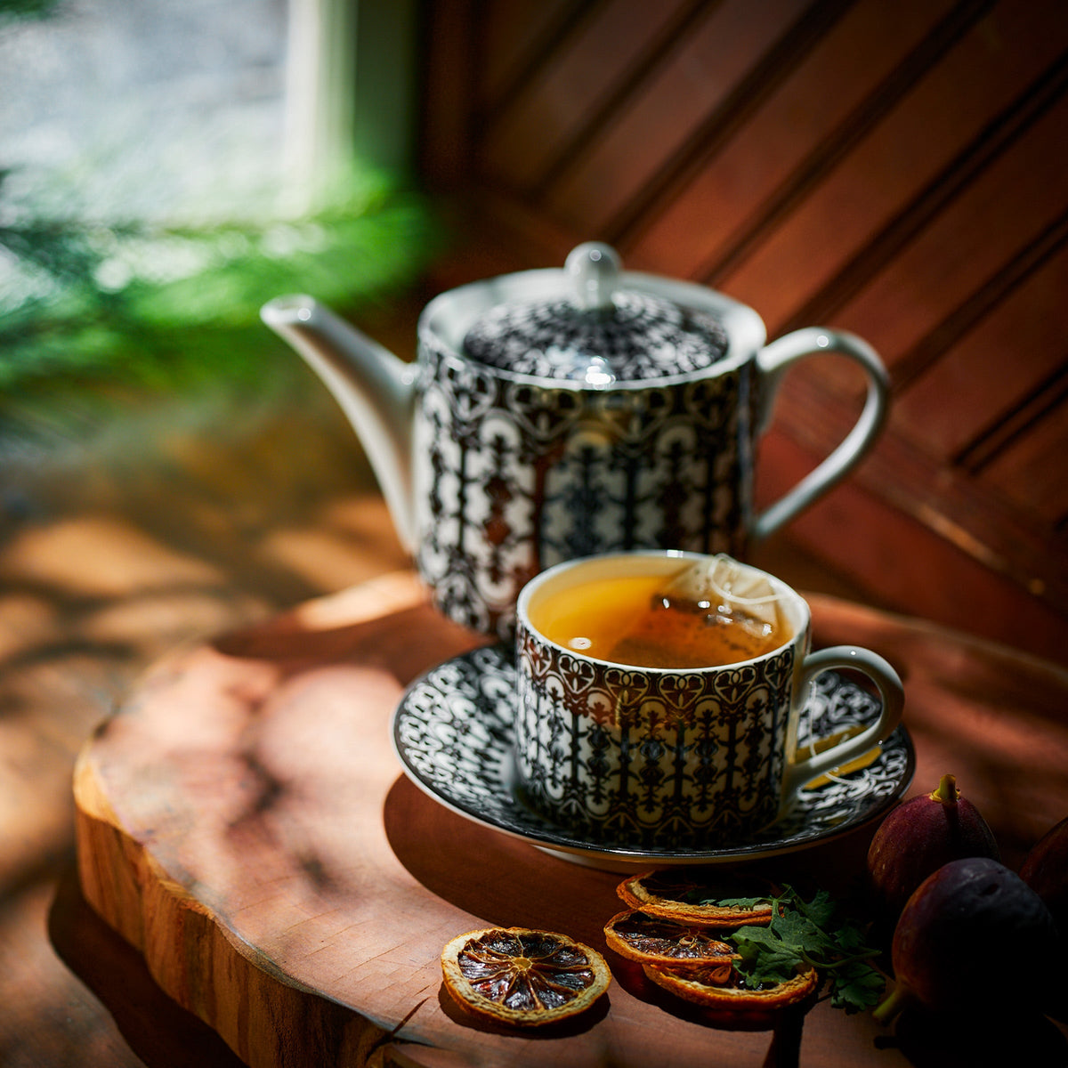 A patterned teapot pours tea into matching Caskata Casablanca Cups &amp; Saucers, Set of 2, placed on a wooden surface with dried citrus slices and figs nearby.