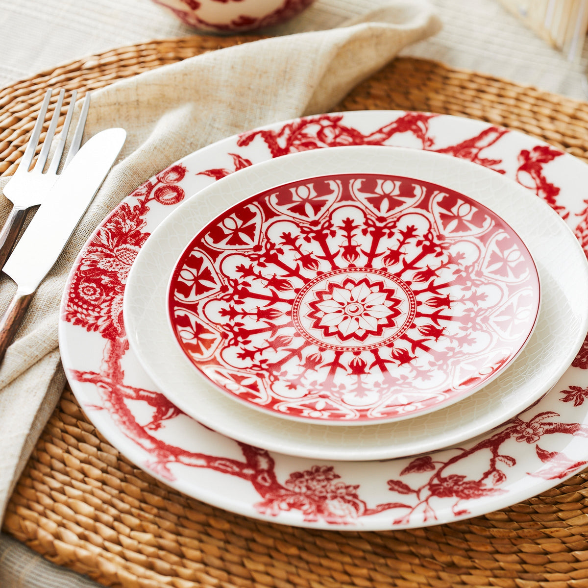 A place setting featuring a white and red patterned **Casablanca Crimson Small Plate by Caskata Artisanal Home** on top of a larger matching Casablanca dinnerware plate, with a fork and knife placed on an ornate scrollwork beige napkin, all arranged on a woven placemat.