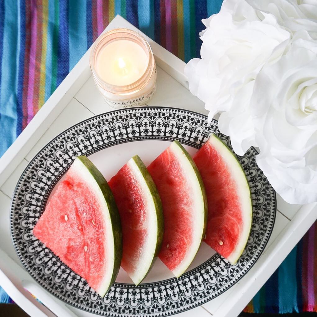 Four watermelon slices arranged on a Caskata Artisanal Home Casablanca Oval Rimmed Platter, placed on a tray with a lit candle and white flowers, all set on a colorful striped background.