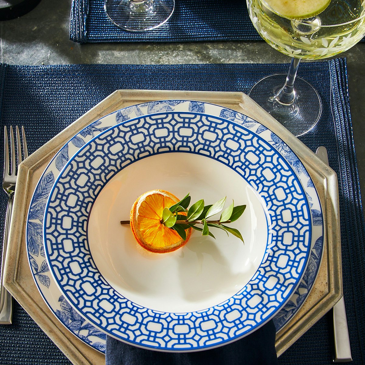 A white Caskata Artisanal Home Arbor Rimmed Charger Plate with a blue geometric pattern holds a slice of orange garnished with a green sprig, set on a blue placemat with a drink in the background.