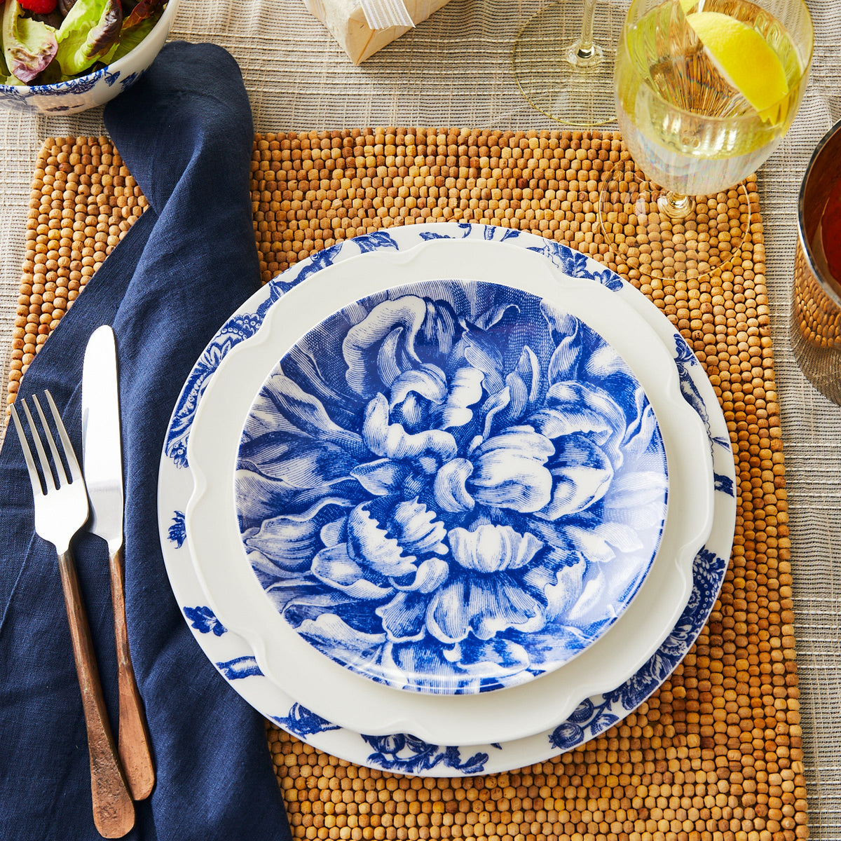 An ornate blue and white Peony Coupe Salad Plate set on a table mat, accompanied by silver utensils, a napkin, and glasses of wine and water.