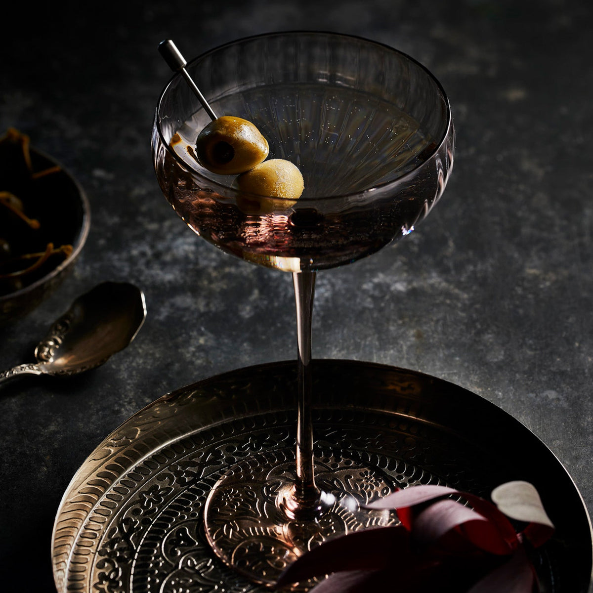 The Quinn Mocha Coupe is a rich brown colored glass, sold as a set, shown here on a silver tray. It holds a martini garnished with olives.