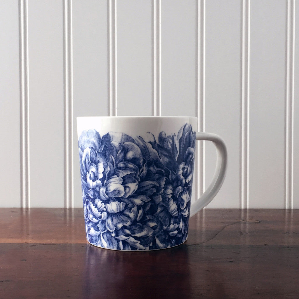 A premium Peony Mug by Caskata Artisanal Home with a blue floral pattern on a wooden table, against a white paneled background.