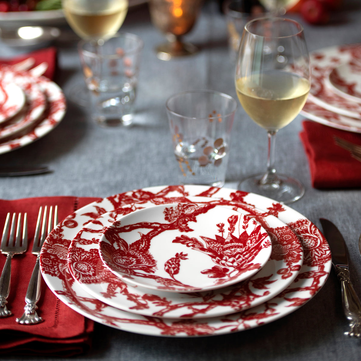 Elegant table setting featuring Caskata Artisanal Home&#39;s Arcadia Crimson Rimmed Salad Plate, silverware on red napkins, and filled wine glasses on a gray tablecloth, evoking an heirloom feel.