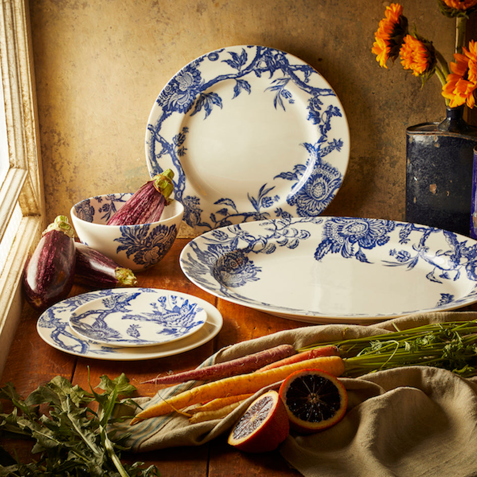 A Caskata Artisanal Home Arcadia Rimmed Dinner Plate featuring blue and white graphic florals is displayed on a wooden table with assorted vegetables, a sliced blood orange, and an arrangement of orange flowers in a vase.
