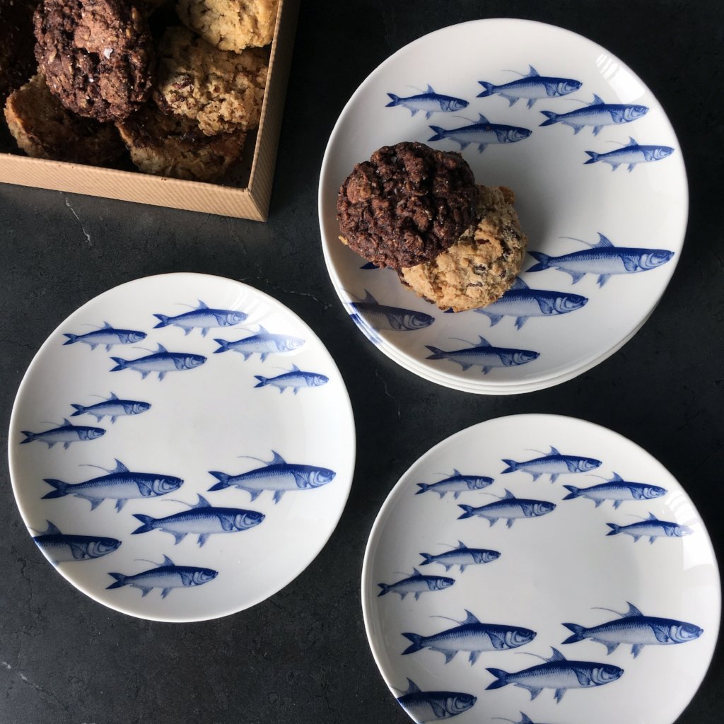 Four white plates adorned with a blue school of fish pattern, one holding a stack of two cookies. In the background, a box of assorted cookies adds to the charm. This heirloom-quality &quot;School of Fish Small Plates&quot; by Caskata Artisanal Home is perfect for any occasion and is also microwave safe.