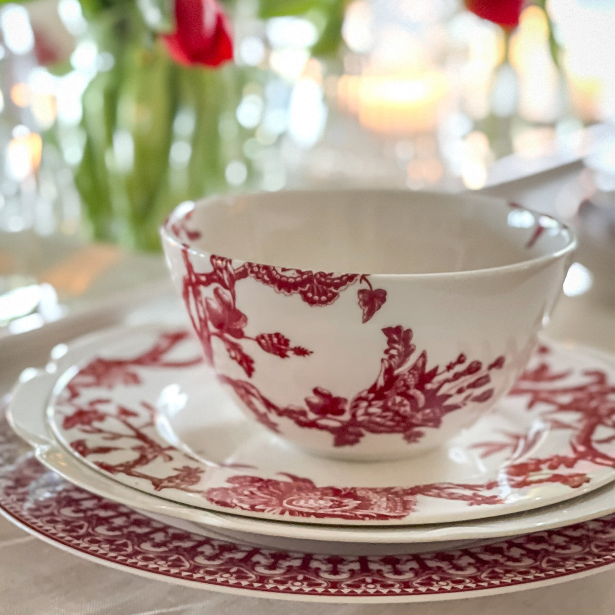 A white porcelain Arcadia Crimson Cereal Bowl by Caskata with red floral patterns, placed on a matching plate, set on a table with a blurred background. Part of the Williamsburg Foundation&#39;s premium porcelain dinnerware collection.