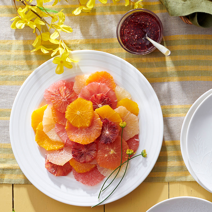 A plate of assorted citrus fruit slices is arranged on a Cambridge Stripe tablecloth beside a jar of jam and a yellow flowering branch, all set atop an elegant high-fired porcelain Caskata Artisanal Home Cambridge Stripe Rimmed Salad Plate.