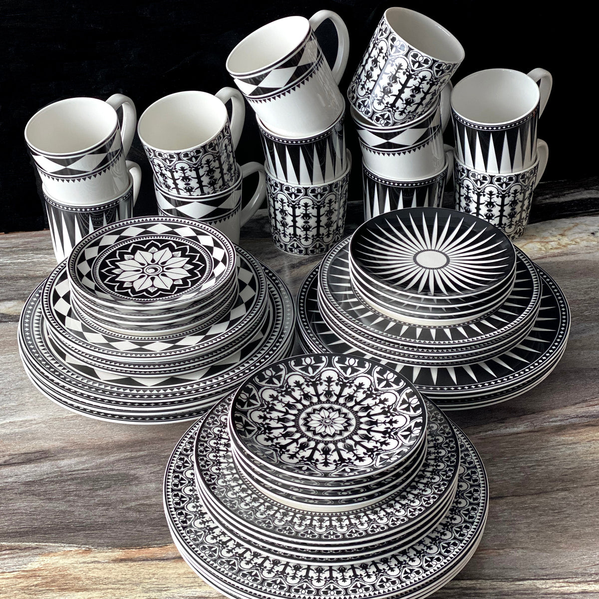 A Casablanca Rimmed Dinner Plate from Caskata Artisanal Home with black and white patterns is arranged on a wooden surface, featuring plates, bowls, and mugs stacked in a neat display. The hand-decorated details add an elegant touch to this high-fired porcelain collection.